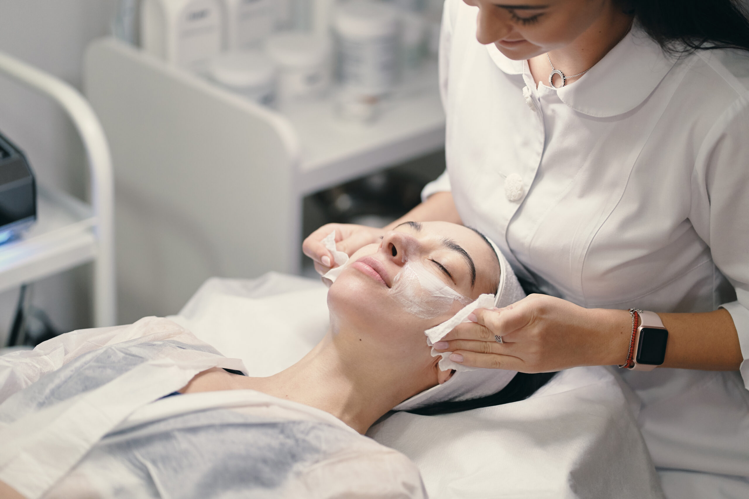 Woman lying during cosmetic procedure while skillful female cosmetologist making facial treatment